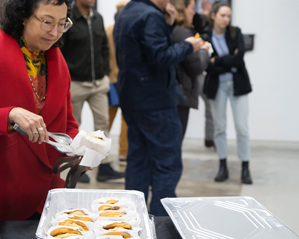 image of a woman wearing a red coat grabbing a burger from a metal tray at the opening reception of Jeremy Fish's exhibition 