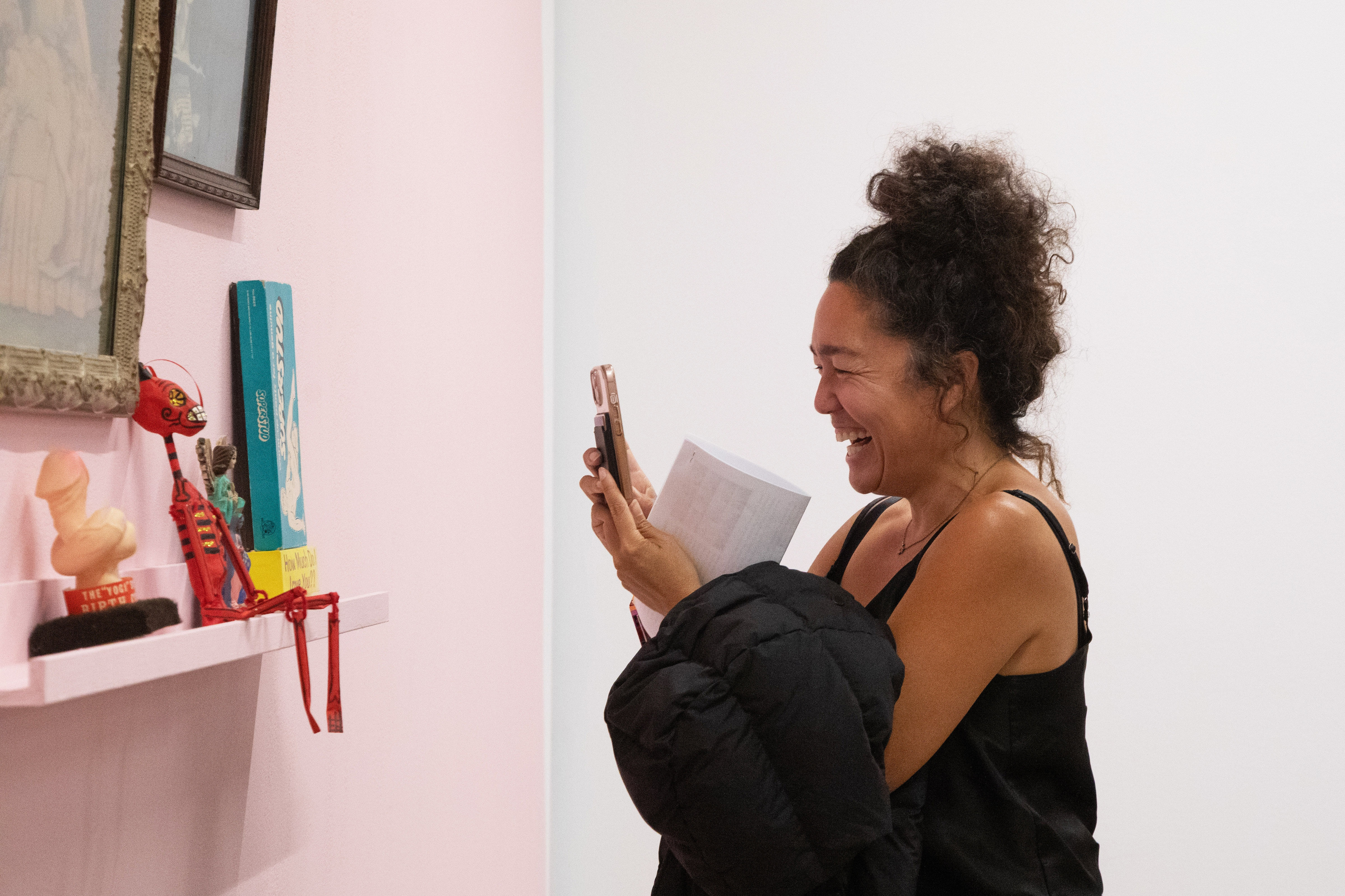A woman takes a photo of an installation on a pink wall while laughing inside an art gallery with fluorescent lighting.