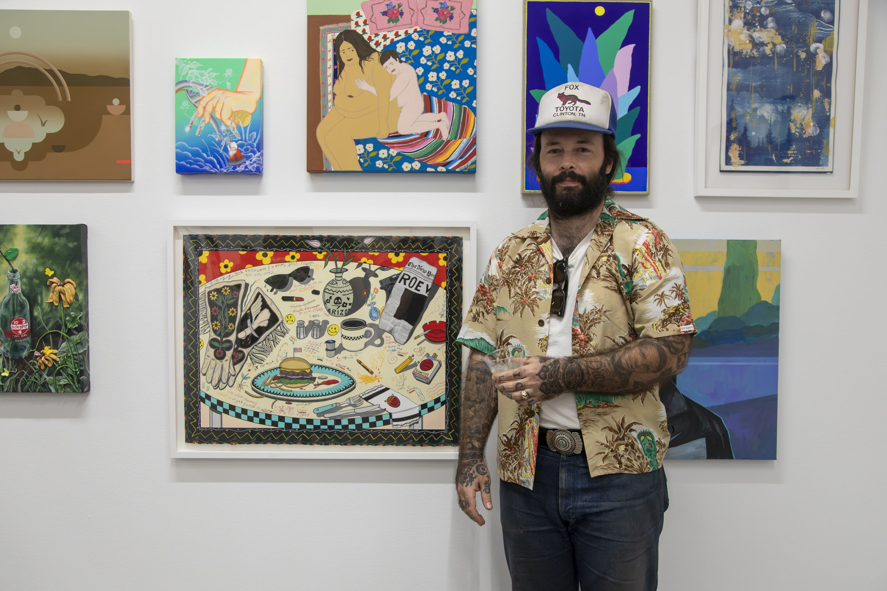 Artist Alex Ziv poses with his artwork against a wall full of other paintings.