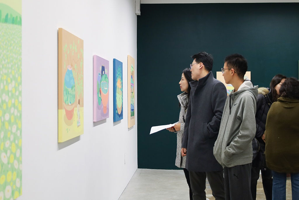 A group of people stand in front of large pastel colored paintings on a white wall, with fluorescent lights above.