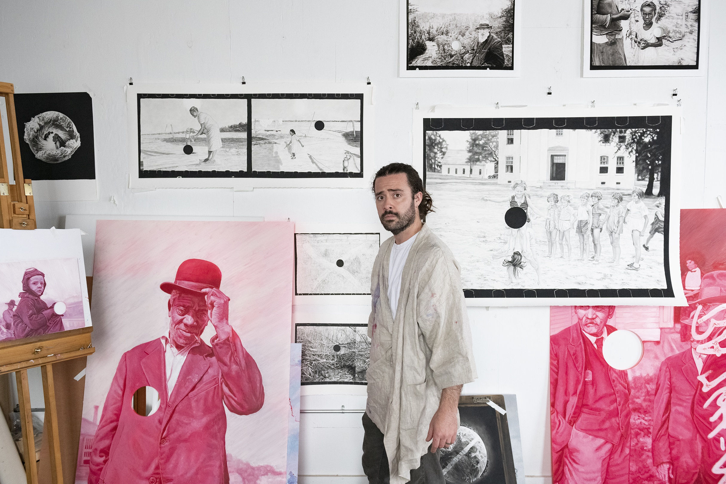 Artist Joel Daniel Phillips stands in his studio among his black and white and red chromatic drawings and paintings. He's wearing a beige robe.