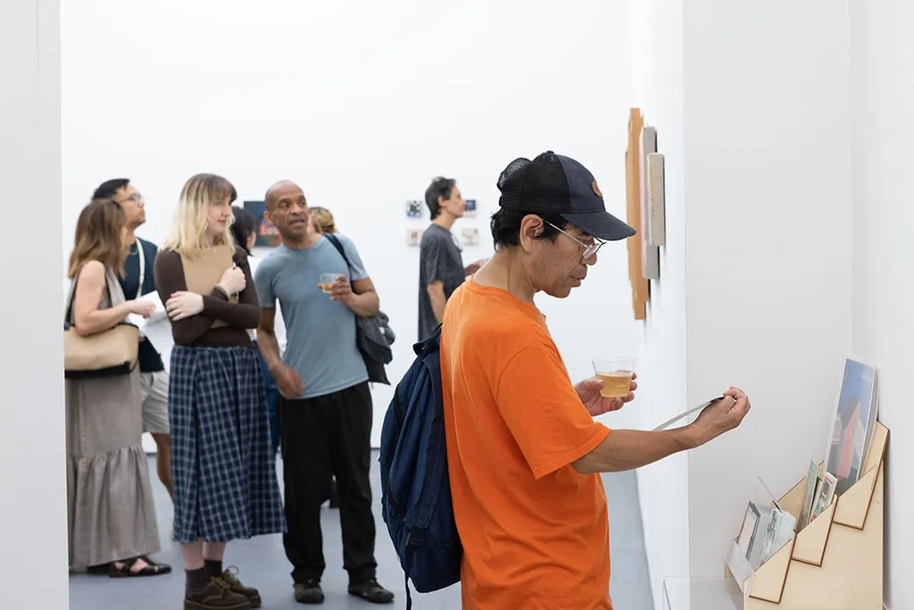 a person looking at a white booklet with a crowd in the background at the opening for Left Unsaid at Hashimoto Contemporary
