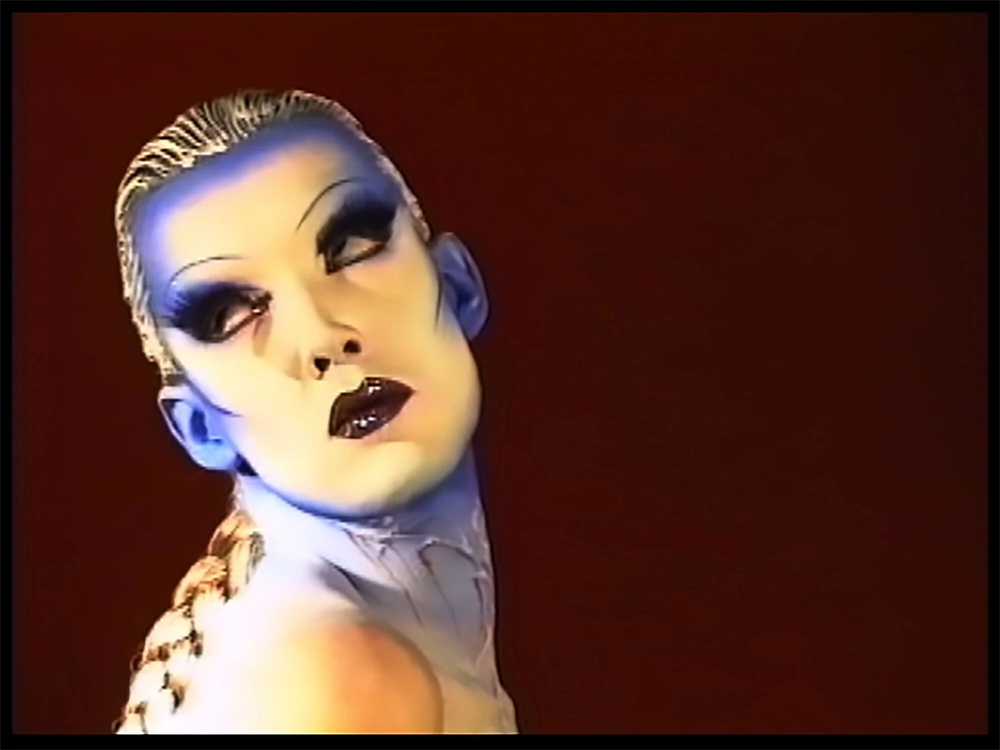 Video still: head shot of a performer in kabuki makeup against a burgundy background