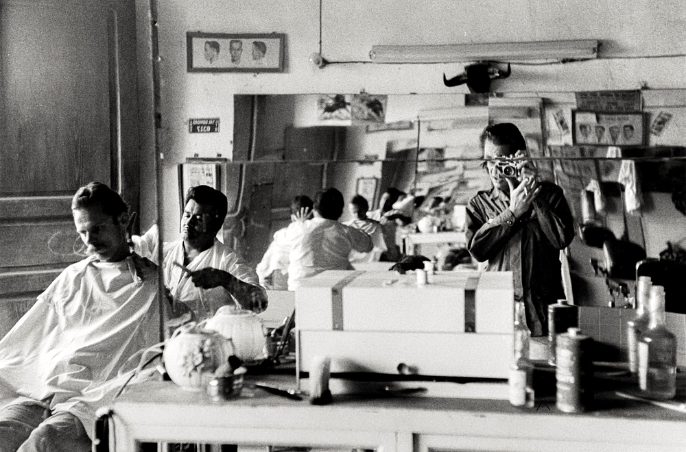 Black and white photo of a photographer taking a picture in the reflection of a mirror in a barber shop