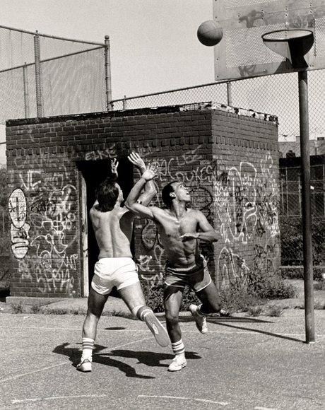 Black and white photo of shirtless two men playing pick-up basketball on an urban court. Both are strking a dramatic poe, as they watch a basketball hat one of them has just shot.