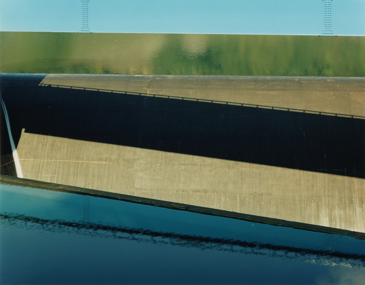 Abstracted color photograph a waterway, dam, and bridge, showing reflections on water.