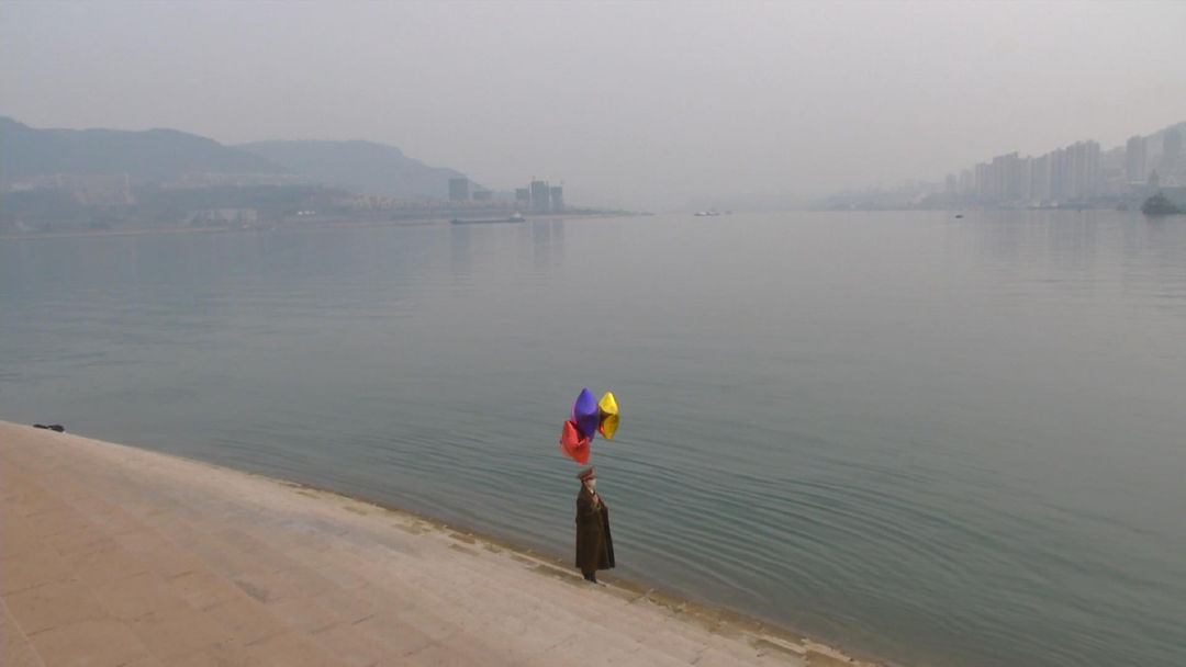 A video still from the Video No.3 out of 7 of Chen Qiulin's The Empty City created in 2012; the still shows the artist Chen Qiulin in a dark green military coat stands on the shore holding balloons of various colors.