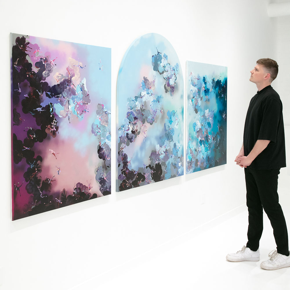 Paul Kuntze Falling Angels exhibition installation view with a person standing in front of a triptych three panel painting with blue, pinks and lavender hues featuring abstract human figures amongst aerosol clouds