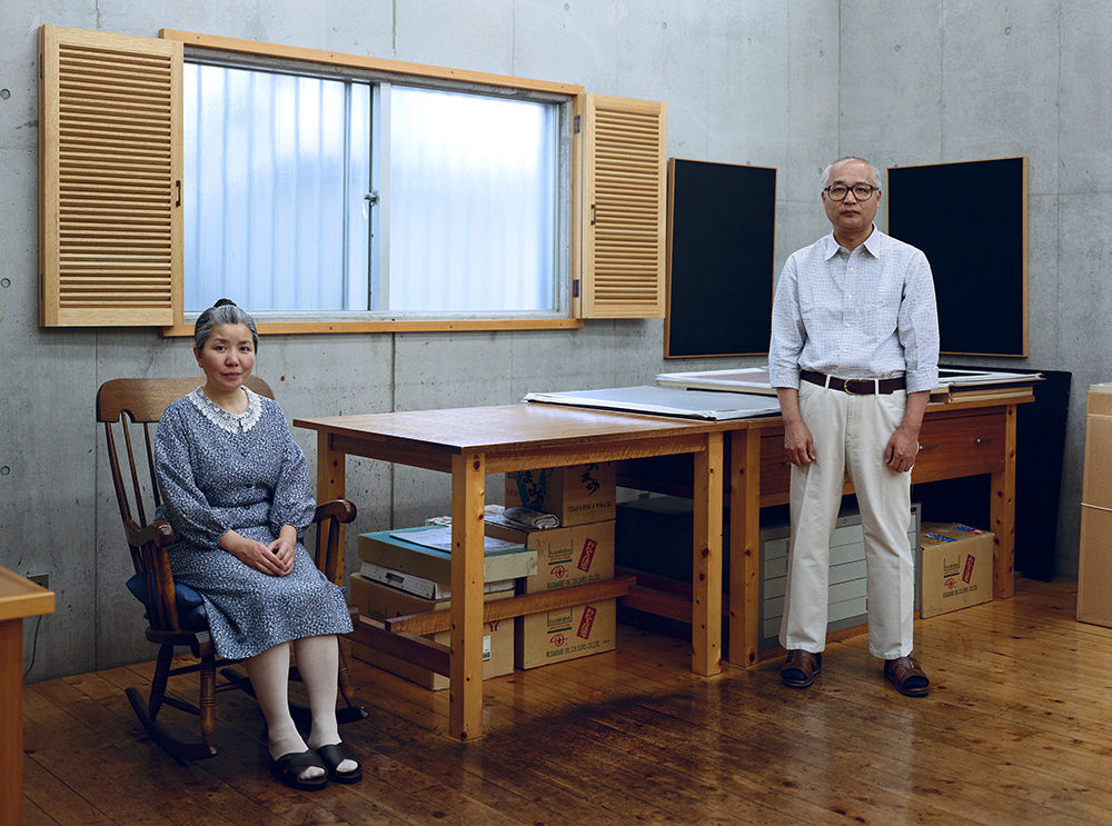 Image of two people gazing into the camera. One person sits in a chair with their hands in their lap. The other stands across the wooden table, looking directly at the camera. The wall is made of cement.