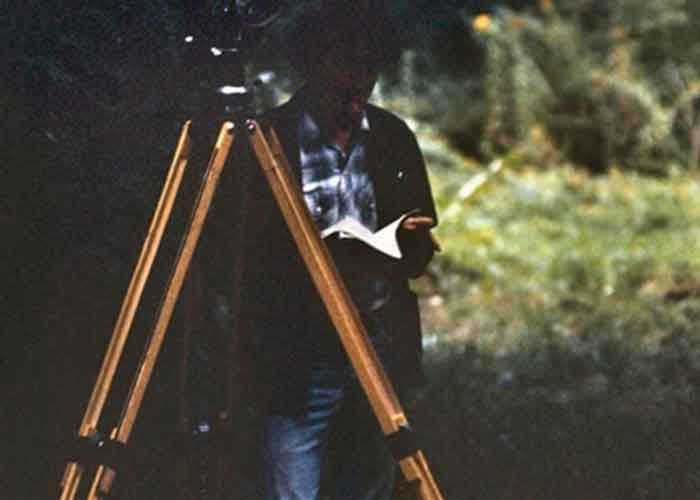 A dark, grainy image of a man standing behind a tripod reading a book. 