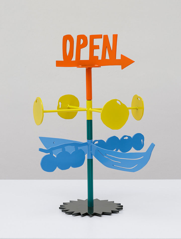 Metal sculpture with the word OPEN on the top and an arrow in orange. Stacked below are four yellow circles on yellow rods and stacked below those are blue berries and bananas on a steal stand.