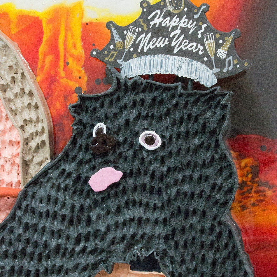 Mixed media hanging sculpture of a woman with a carrot in her mouth holding a small black dog with a Happy New Year paper crown on his head and a pretzel and a carrot in his hands. In the background an atomic bomb is going off.