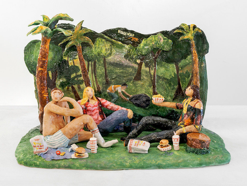 Scenic sculpture of a group of people sitting in a park under palm trees eating in n out on the grass.