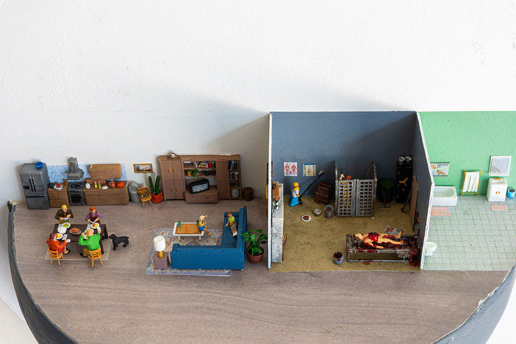 miniature diorama by Abigail Goldman of the interior of house with three rooms, one in which a family is eating dinner and another with people chained and bleeding