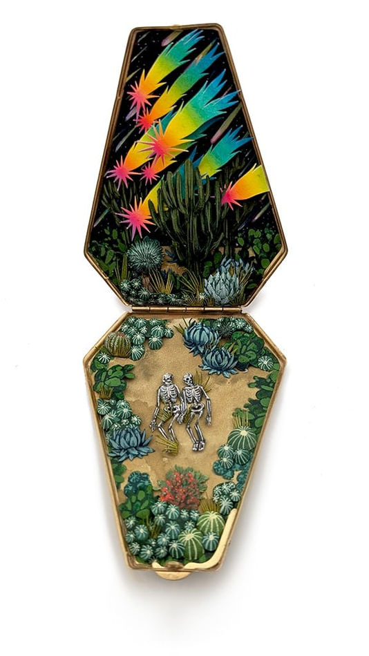 hand-cut watercolor collage in antique cosmetic compact by Shannon Taylor of two skeletons laying down on the ground with cactus surrounding them and colorful stars above them