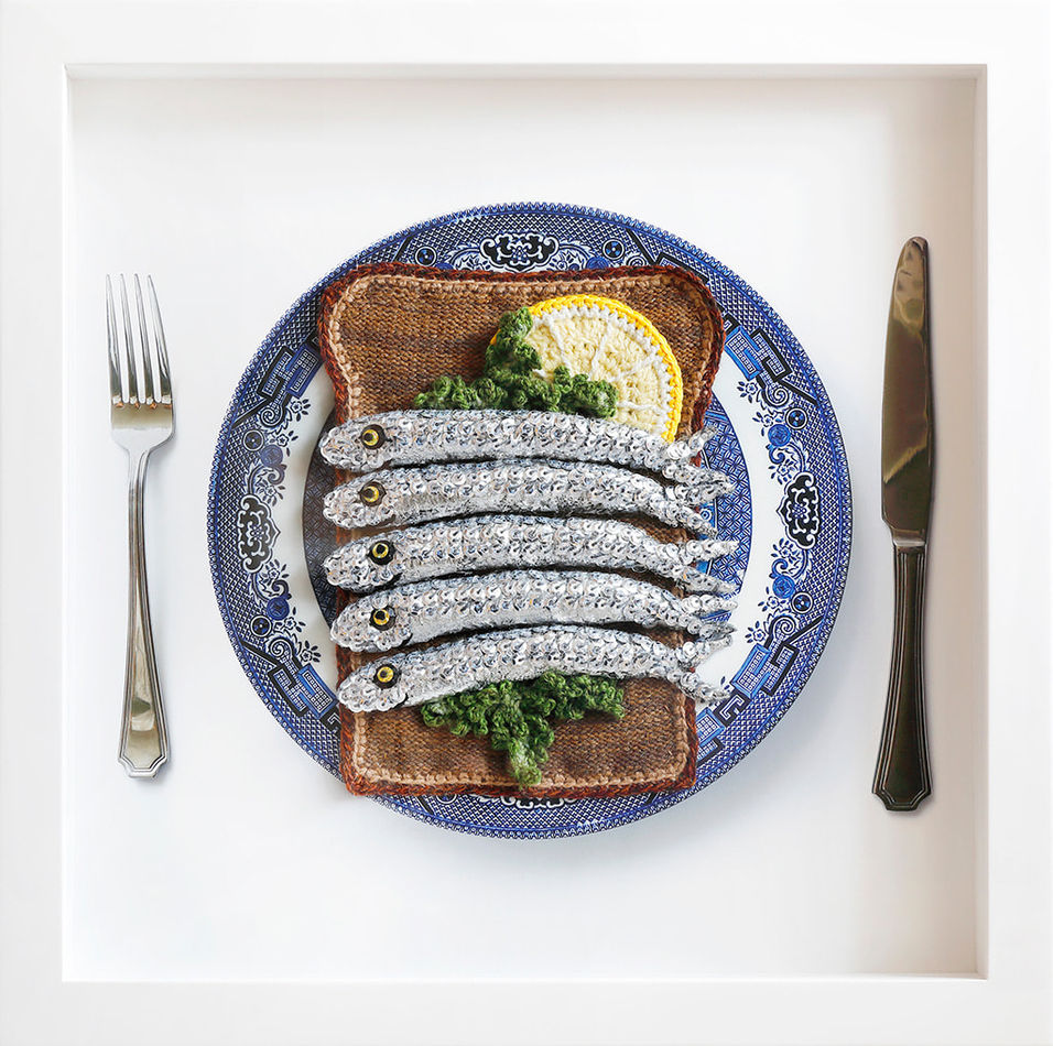 Soft sculpture of crocheted sardines on a bed of lettuce on top of a piece of toast on a blue plate with a fork and knife