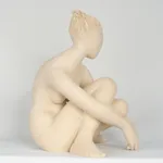 Seated white ceramic figure is looking over her left shoulder with arms draped around her knees which are tucked together.