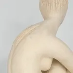 Seated white ceramic figure is looking over her left shoulder with arms draped around her knees which are tucked together.