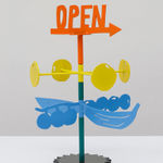 Metal sculpture with the word OPEN on the top and an arrow in orange. Stacked below are four yellow circles on yellow rods and stacked below those are blue berries and bananas on a steal stand.