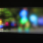 Still image of a rectangular piece with multi-colored LEDs behind angled Plexiglass that display scenes of people running To the right side the scene is burry due to the Plexi being farther away while at the left is it more pixelated due to the Plexiglass being closer