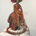 An image of a victorian doll in a vintage silk wedding sari with gold thread brocade, vintage brass claw furniture cap, steel, copper leaf, copper nails, wood, silk, cotton, sequins, gourd, polyester threads, wax, cowry shells, eyelashes, silk trim, pheasant feathers on felt hat plates.