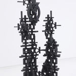 Black steel floor sculpture in two columns, one taller than the other. Columns are composed of interlocking cultural symbols such as ankhs and double happiness