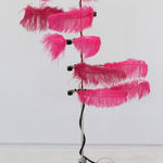 Tall silver pole with five large neon pink feathers on either side that move in all sorts of patterns. There are red and black cords wrapped around pole leading to two black poles at the base spread out for support.