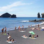Photograph by Massimo Vitali showing beachgoers relaxing on a black sand beach with rocky sea stacks rising from the blue ocean in the background.