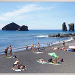 Photograph by Massimo Vitali showing beachgoers relaxing on a black sand beach with rocky sea stacks rising from the blue ocean in the background.