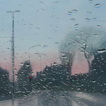 A wet, city road leading to silhouettes of buildings with a pink and light blue sky backdrop, seen through a glass screen with water droplets scattered across it. By artist Esther Nienhuis.