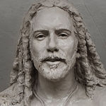 A clay model close-up of the statue during the sculpting process, showcasing the intricate details of Jesus' face and hair before the casting stage, with a neutral gray background.