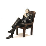 A resin sculpture titled Chairman depicting a seated male figure in a suit. The man is portrayed in a thoughtful pose, leaning back slightly in a high-backed chair with his legs crossed and hands clasped together. The figure’s head and hands have chrome finishes, contrasting with the darker patina of the suit and chair. The piece exudes authority and contemplation.
