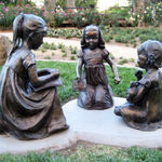 Bronze sculptures of three young girls seated in a circle on a grass-covered garden pathway. One girl holds an open book, another clutches a teddy bear, and the third engages attentively, creating a serene and playful storytelling scene. The setting features vibrant rose bushes and a tranquil garden backdrop.
