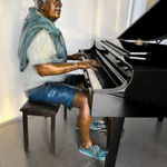 Bronze sculpture of an elderly man seated at a grand piano, playing with a serene and focused expression. The figure is dressed casually in shorts, a t-shirt, and a sweater draped over his shoulders, with vibrant color accents highlighting his clothing and shoes. The artwork captures a moment of musical engagement and nostalgia in a modern indoor setting.