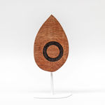 Wooden sculpture of a tear drop shaped wooden piece with a black circle on a white steal stand