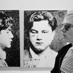 Black and white photograph of Andy Warhol in profile in front of a screen print of a man’s mug shot