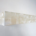 Detail of a row of white iridescent cubes made of acrylic paint on MDF with a pale pink textured strip at its center is mounted on a white wall.