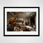 Framed photograph in black wood of an artist, Gilda Pervin, standing amidst an array of work desks, art materials, and books in a loft setting