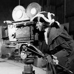 Black and white photograph of Jean Cocteau dressed in the style of an 18th century nobleman wearing a hat, looking to the left into a film camera