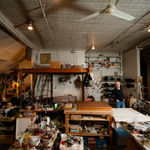 An artist, Gilda Pervin, standing amidst an array of work desks, art materials, and books in a loft setting