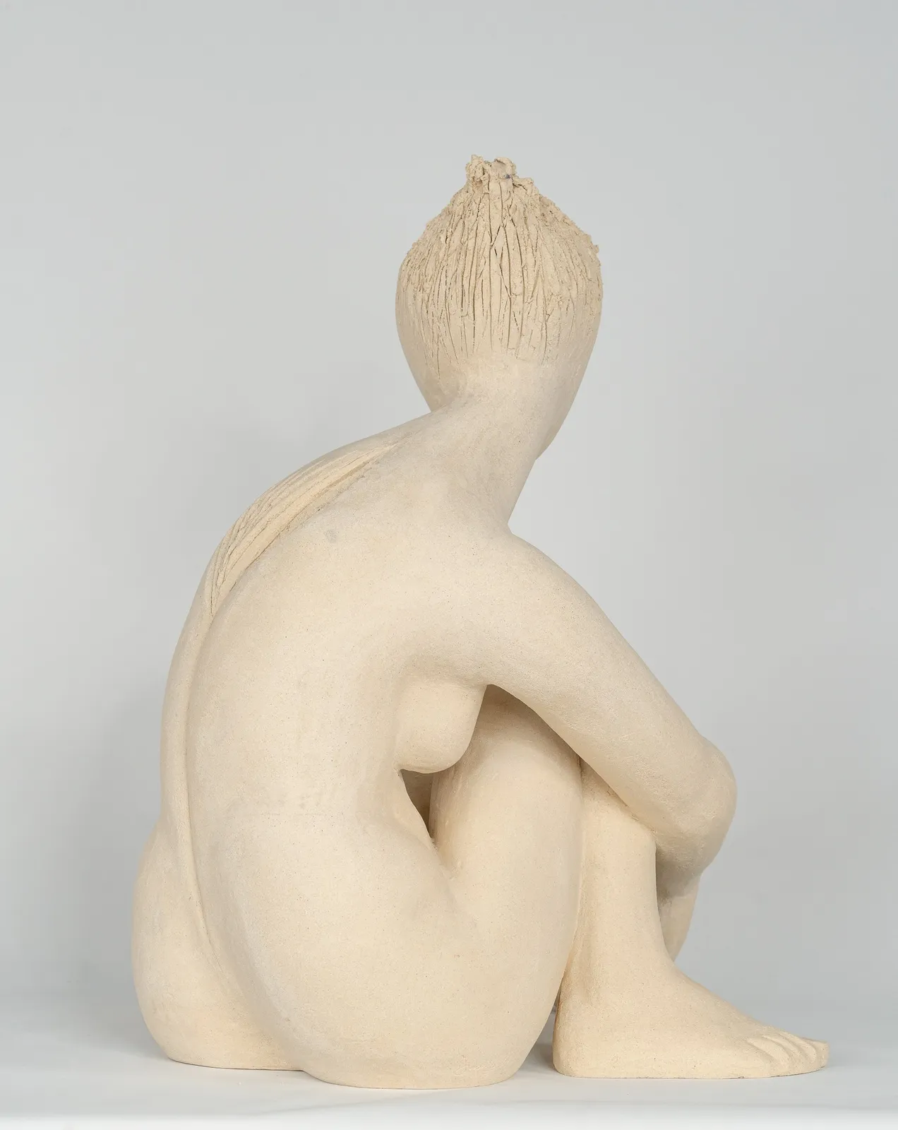 Seated white ceramic figure is looking over her left shoulder with arms draped around her knees which are tucked together.