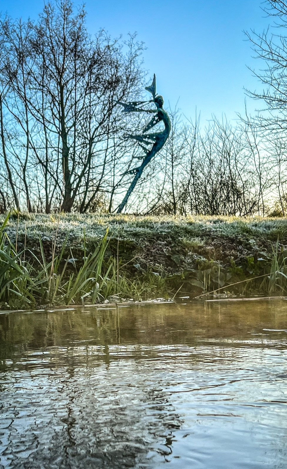 A monumental bronze sculpture of a winged female stands in front of trees at a Dorset sculpture park, Sculpture by the Lakes, lake in foreground