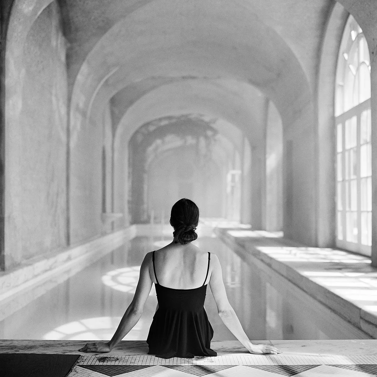 Black and white photograph of woman sitting on edge of indoor pool