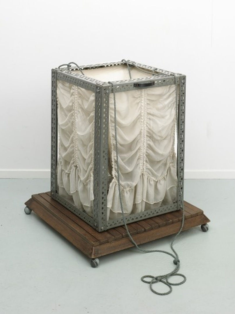 A glass box with metal frames sitting on a small wooden platform; each side of the box is covered in white curtains.