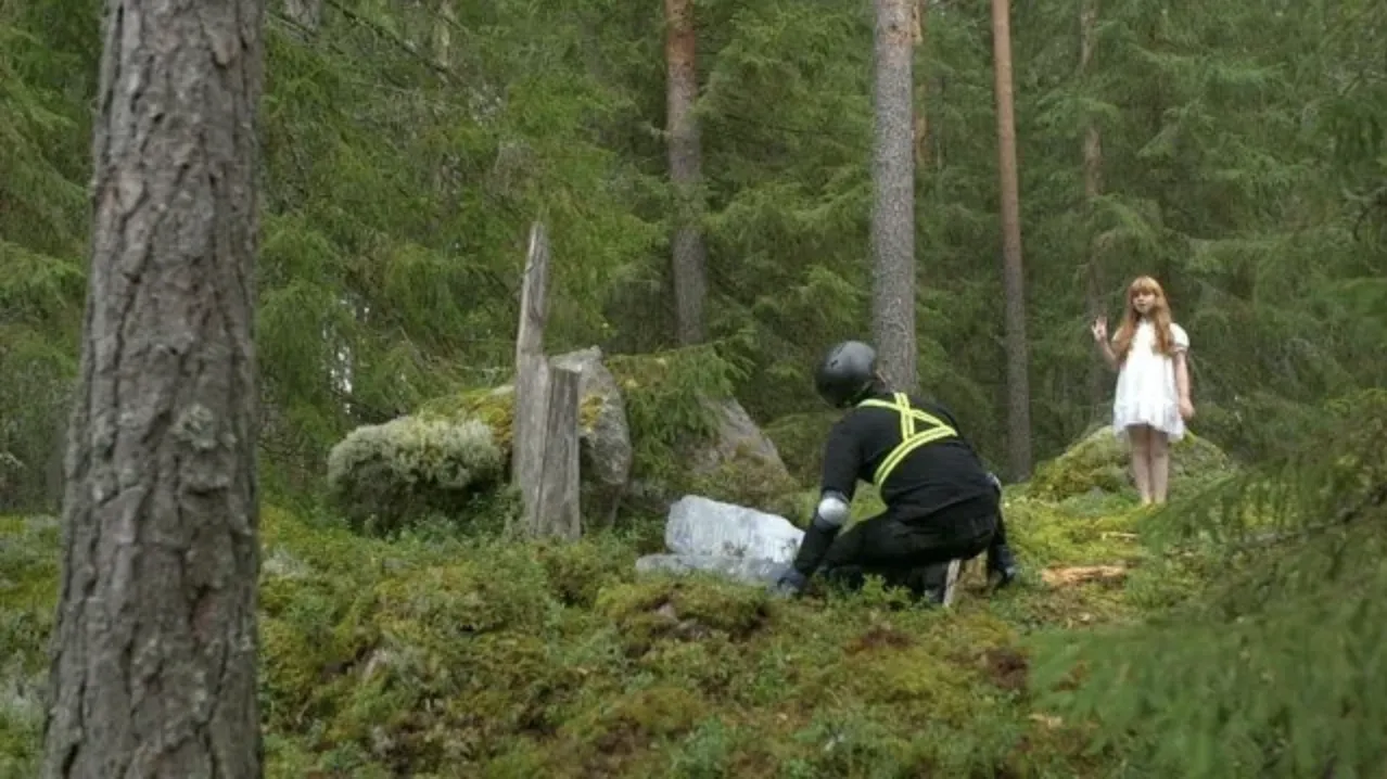 A still from a 4K video depicting a scene in a lush forest where a young girl in a white dress waves to an adult who appears to be some kind of forest ranger.
