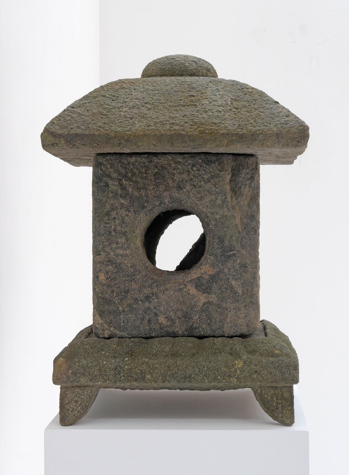 Weathered stone lantern with square base and feet, square body with hole in the center, and domed topper.