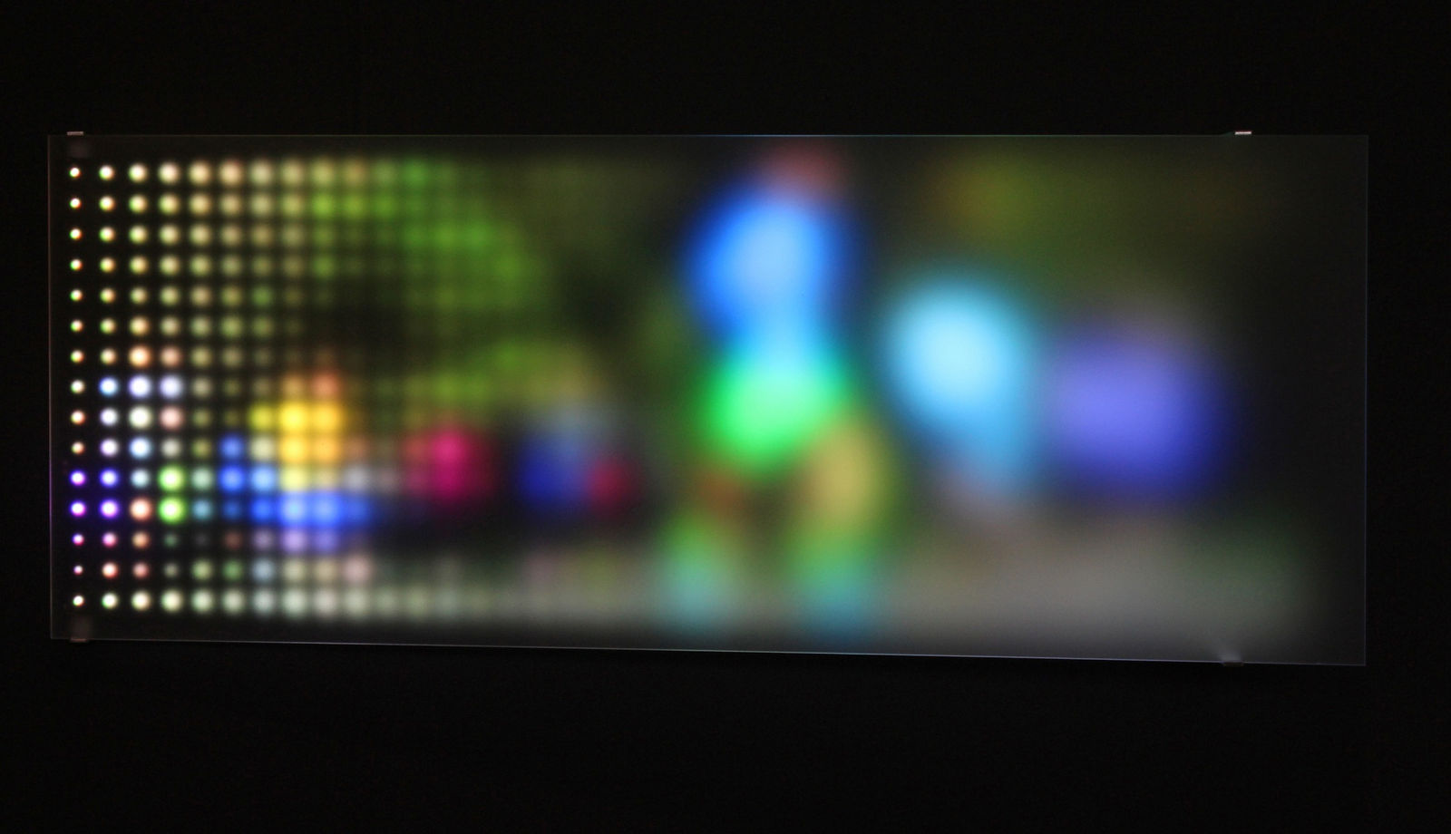 Still image of a rectangular piece with multi-colored LEDs behind angled Plexiglass that display scenes of people running To the right side the scene is burry due to the Plexi being farther away while at the left is it more pixelated due to the Plexiglass being closer
