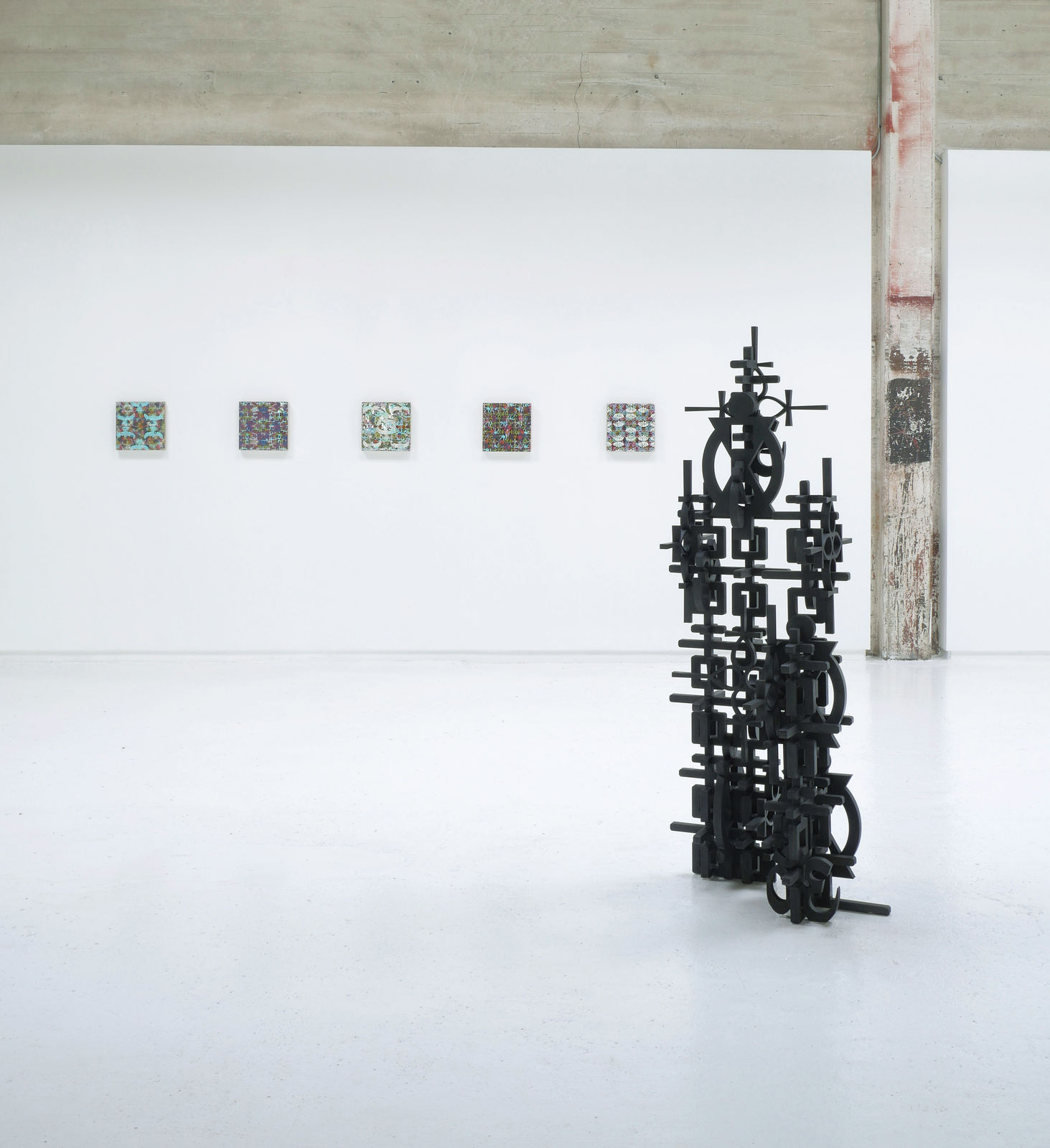 Installation view of Totem with 5 square panel paintings hanging in a horizontal line on the wall behind the sculpture