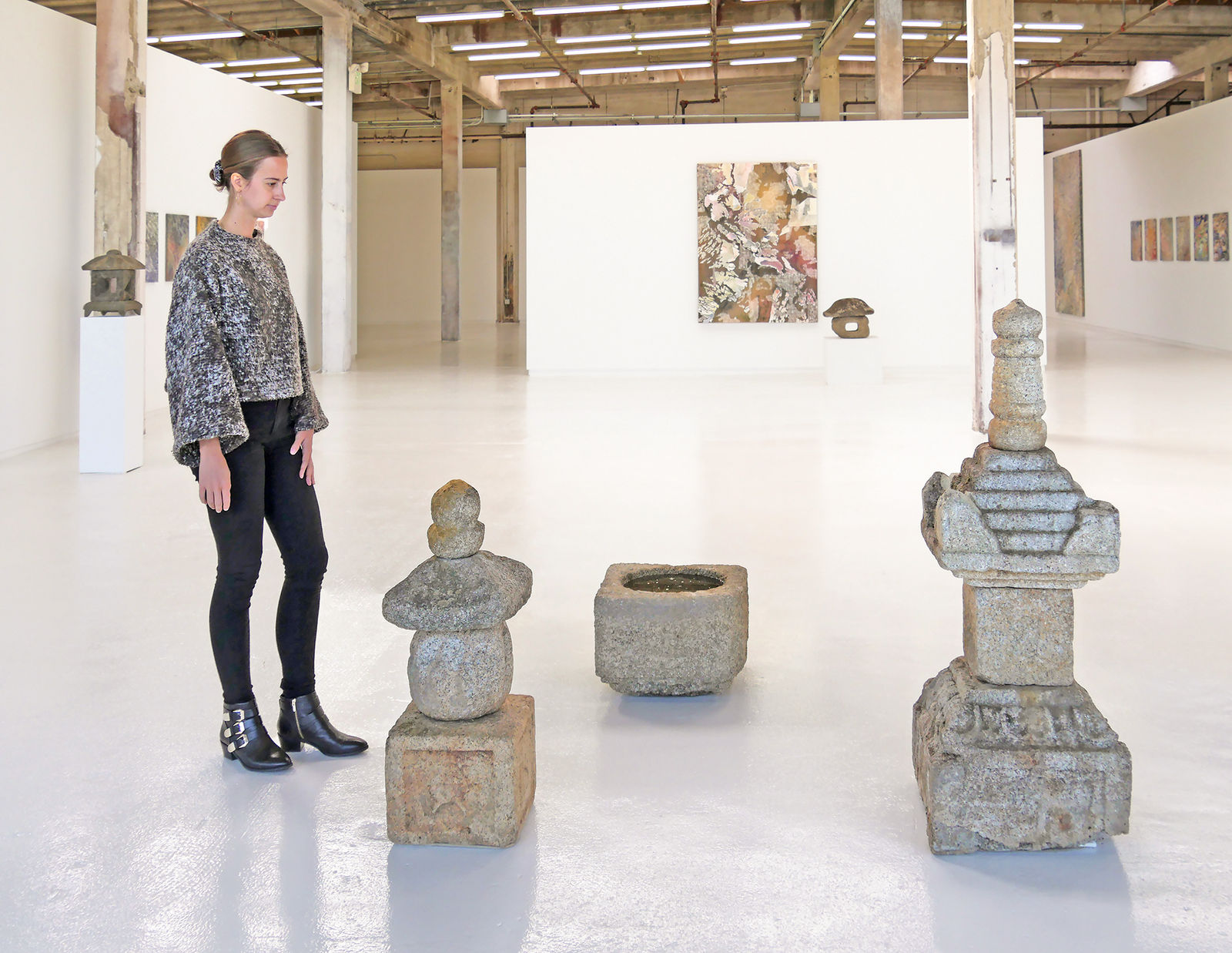 Woman standing with assortment of Japanese stone sculptures in gallery with colorful paintings on back wall.