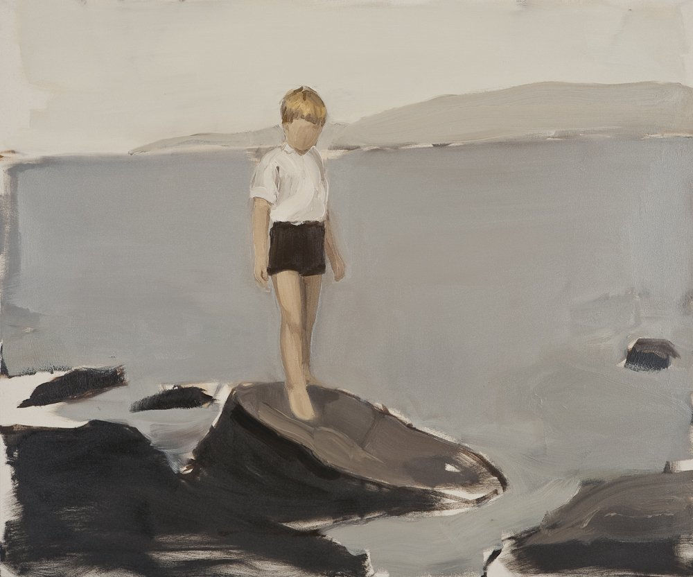 Young faceless blonde boy in white shirt and black shorts stands on rocks at the shore of a lake with water, mountains and sky in the background.